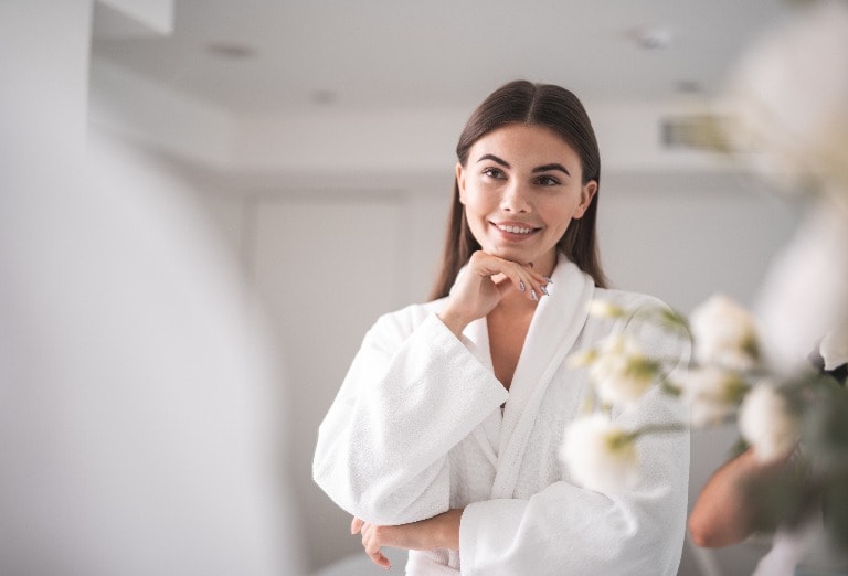 I am so beautiful. Portrait of optimistic lady looking at mirror while touching chin with arm. She standing in room