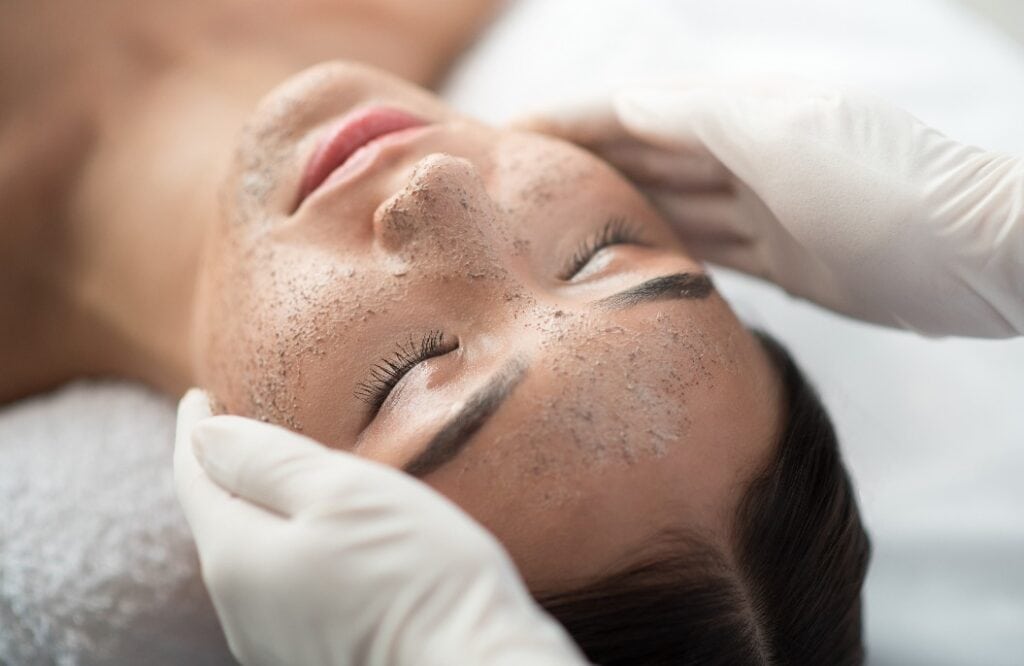 Close up of young woman getting facial skin treatment at spa. Cosmetician hands applying peeling mask on her face