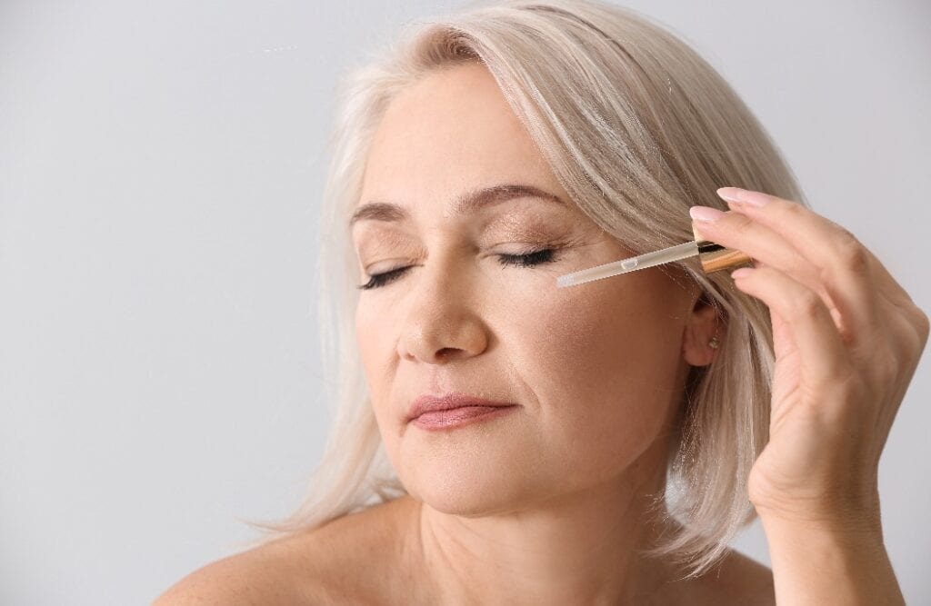 Mature woman applying face serum on light background